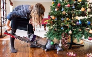 Kuvassa nainen imuroi koristellun joulukuusen alle pudonneita neulasia.