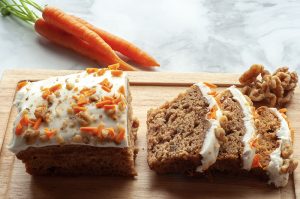 Ylhäältä kuvattuna leikkuulaudalla porkkanakakku, josta on leikattu kolme viipaletta. Taustalla kolme porkkanaa naatteineen marmoripintaisella pöydällä. Leikkuulaudalla on myös pieni määrä pähkinöitä.
