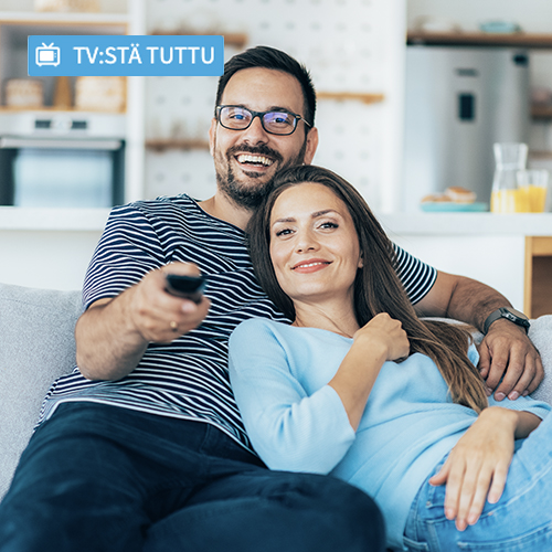 Hymyilevä pariskunta istuu sohvalla ja katsoo televisiota kotonaan, mies pitelee kaukosäädintä.