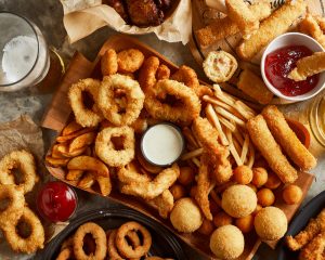 Ylhäältä kuvattuna tarjottimia, joilla on ranskanperunoita, friteerattuja sipulirenkaita, mozzarellatikkuja ja muita rasvapaistettuja ruokia sekä niille tarkoitettuja dippikastikkeita ja olutlasi.