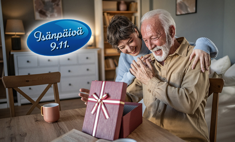 Iloisesti yllättynyt harmaapartainen mies avaa lahjarasiaa ruokapöydän ääressä. Häntä onnitteleva nainen halaa takaapäin.