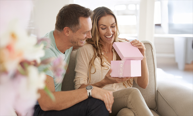 Keski-ikäinen pariskunta istuu sohvalla, nainen avaa vaaleanpunaista lahjarasiaa iloisesti yllättynyt ilme kasvoillaan. Mies hymyilee vieressä. Kuvan etualalla näkyy epäterävänä kukkakimppu.
