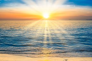 Merenranta auringon laskiessa horisonttiin. Meren aallot ovat lempeitä, taivaan pilvet hehkuvat oranssin ja sinisen sävyissä.