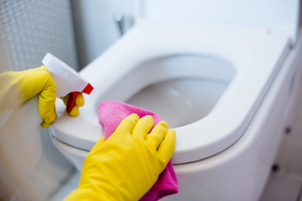 Kädet keltaisissa suojakäsineissä puhdistavat wc-istuinta suihkepullolla ja liinalla.